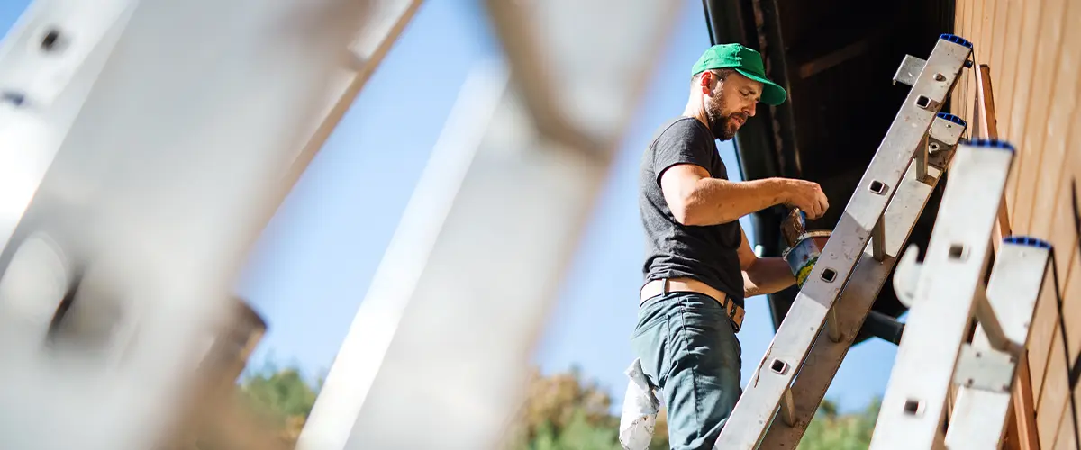 Professional house painter on ladder painting exterior siding with brush. Home improvement and exterior renovation services.